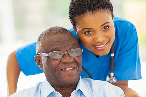 Young nursing in blue scrubs leaning over the shoulder of patient in wheelchair smiling